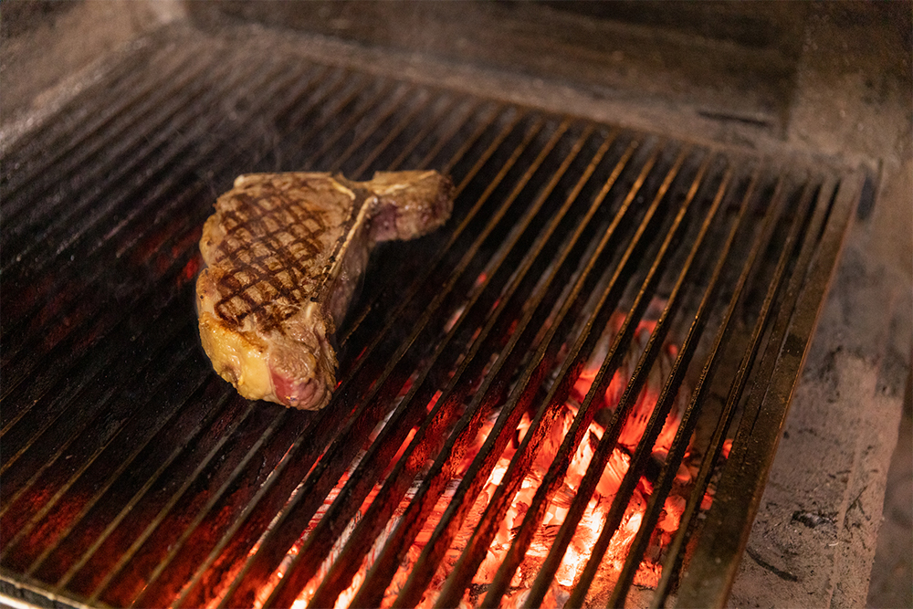 幅3cmのステンレス鋼が0.5cm間隔で連なる焼き網は炉床から高さ10cmの位置に。「強火の近火」で下の熾火から当たる熱だけで肉を焼く。真上のダクトが強力に排気するため、熱はこもらず、放熱性が高い。「輻射熱は要らない」「肉に必要なのは下からの拡散された強い熱」とする渡邊シェフの考え方を実現する構造。