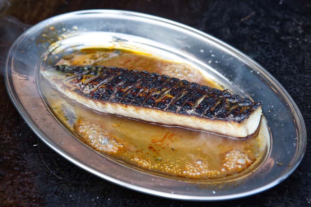 (写真)新鮮なサバは強い炎で皮目をさっと焦がしてからソースと一緒にステンレス皿にのせ、熾火の上で熱して香りを立てる。