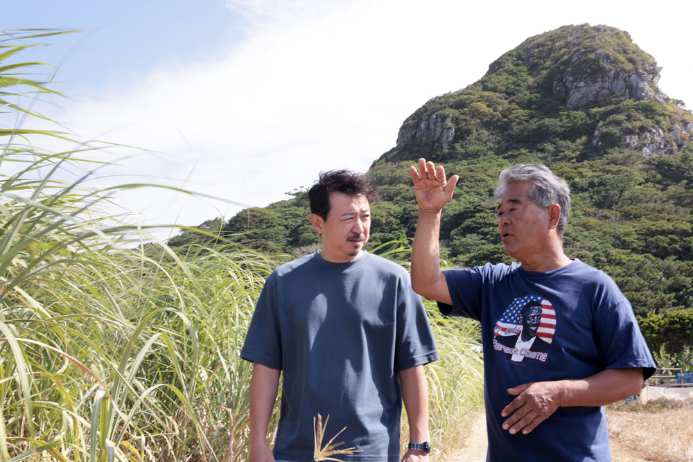 サトウキビ農家の知念和幸さん(右)、宮﨑慎太郎シェフ(左)。後ろにそびえるのは、伊江島のシンボル・城山(ぐすくやま)。